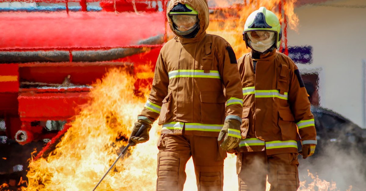Two firefighters wearing helmets, face masks, and flame-resistant uniforms walk together in front of a fire.