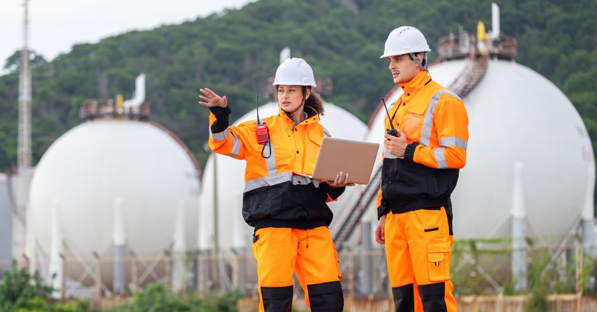 Two workers wearing bright orange and black flame-resistant coveralls and hard hats while talking at an oil site.