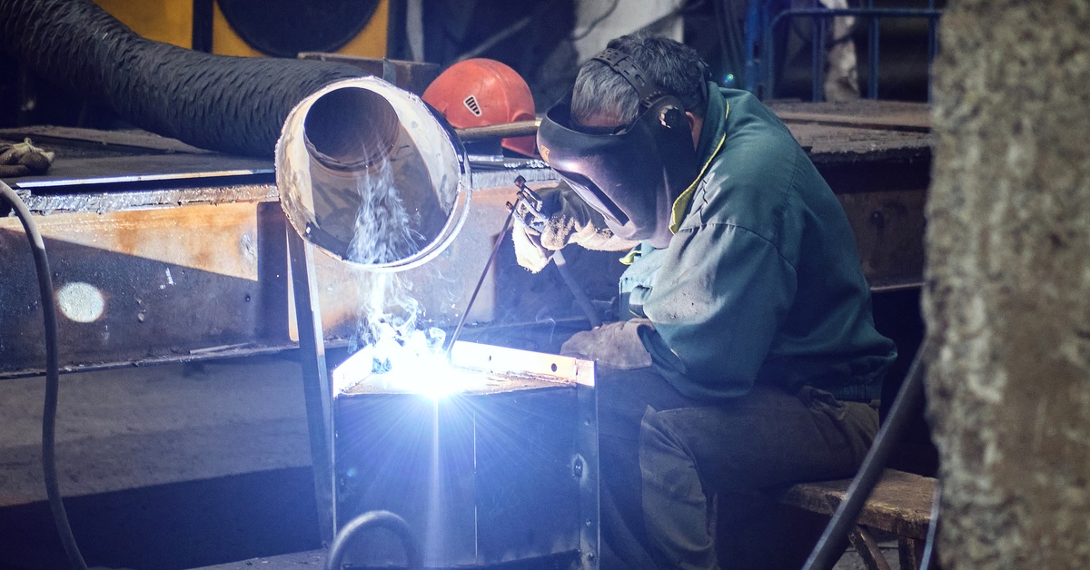 A man is sitting and using a welding machine. He is wearing a protective mask and is wearing gloves.