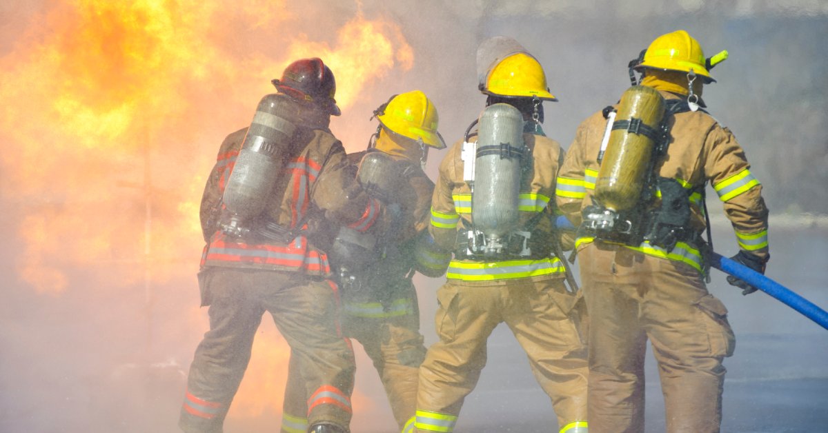 Four firefighters in a training exercise are holding a hose, which is blasting water at flames. They are wearing fire-resistant uniforms.