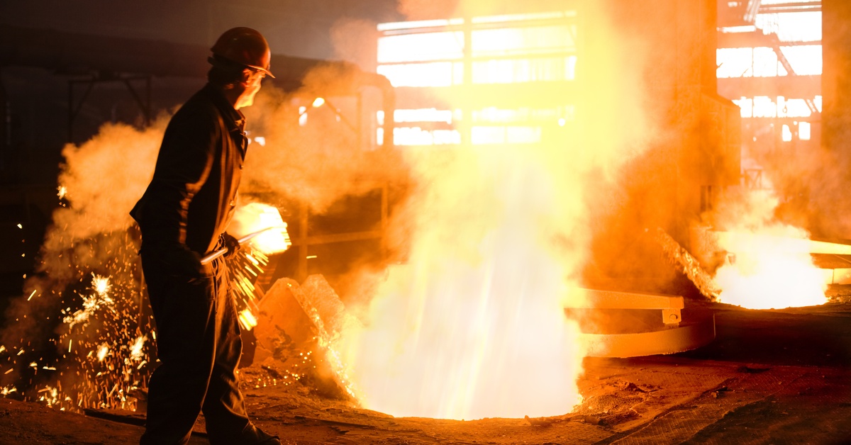 A man standing next to a vat of molten material. The man is holding a shovel and is wearing a hardhat and gloves.