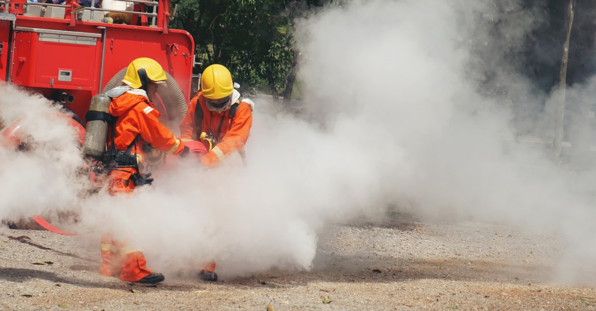 Two people in orange clothing are standing next to a cloud of smoke. There is a red vehicle behind them.