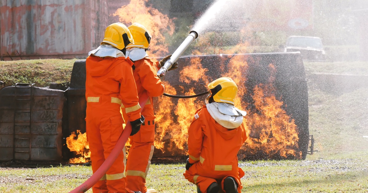 A group of three people are holding a firehose. They are spraying water at a distant fire. They are all wearing orange.