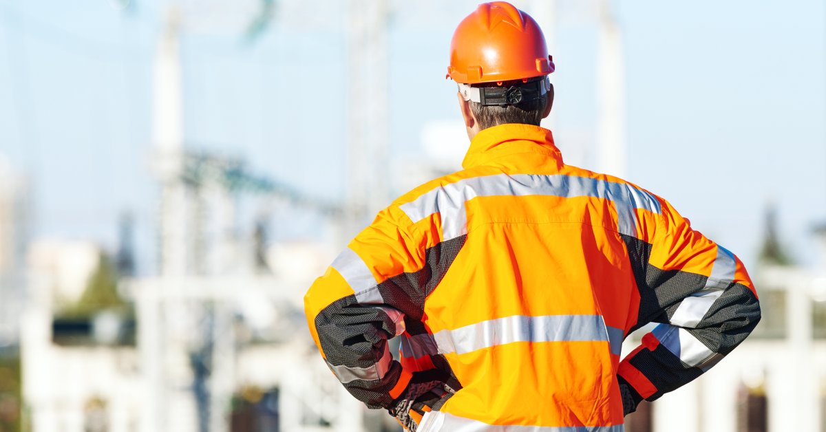 A man in an orange protective work jacket and a hard hat standing with his hands on his hips outside on a job site.