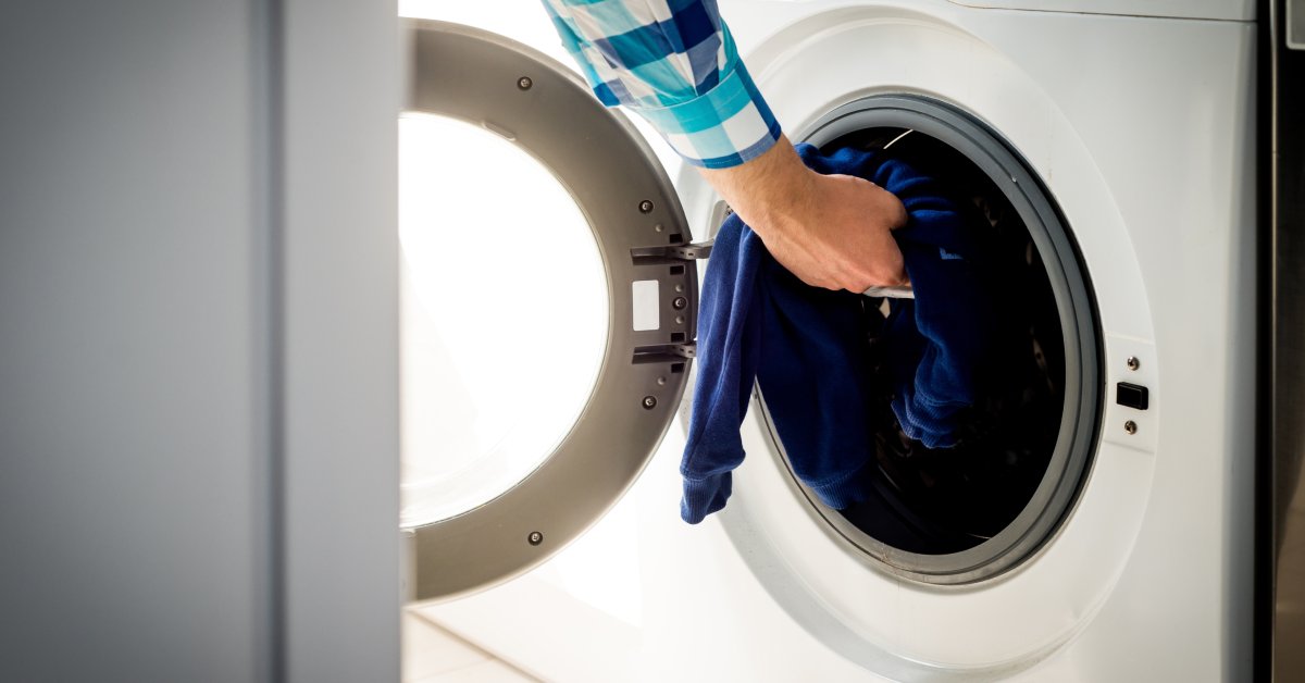 A person reaches into the front opening of a white washing machine, tightly holding blue clothing inside.