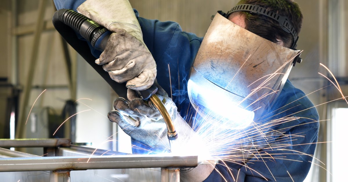 A welder in blue protective clothes, gloves, and a welding mask uses his torching tool on metal pieces.