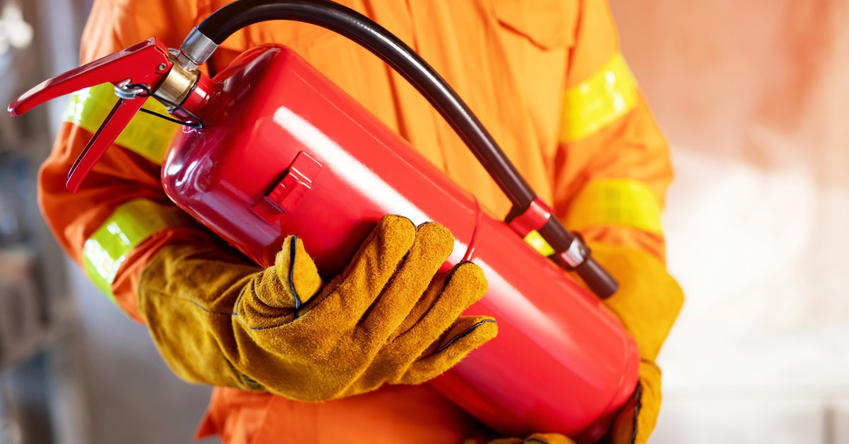 A firefighter in a full orange fire protective outfit holds a red fire extinguisher with both gloved hands.