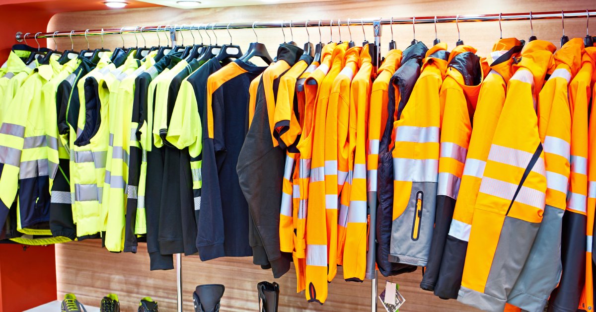 A variety of different yellow and orange industrial workplace clothes hang on a rack with shoes underneath.