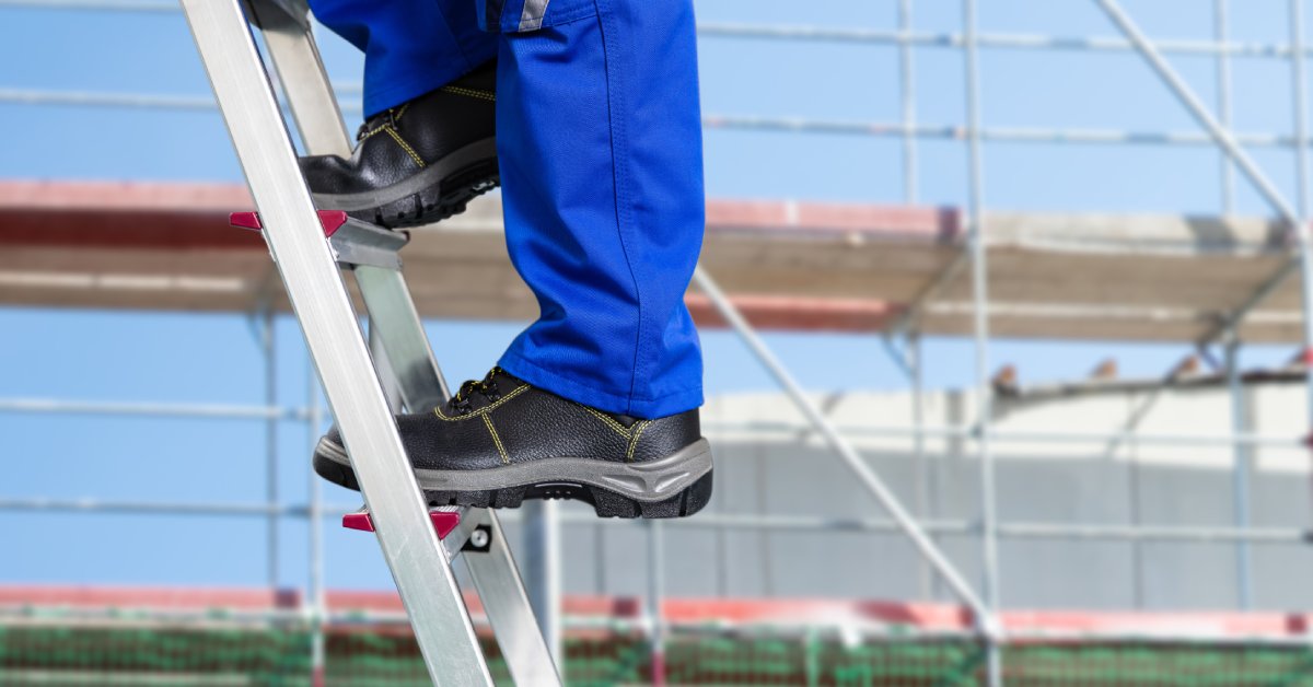 A worker wearing strong black boots walks up a metal ladder in front of a building under construction.