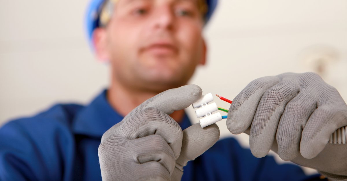 An electrician wearing a hard hat and gray gloves threads three wires into separate sections in a piece of plastic.