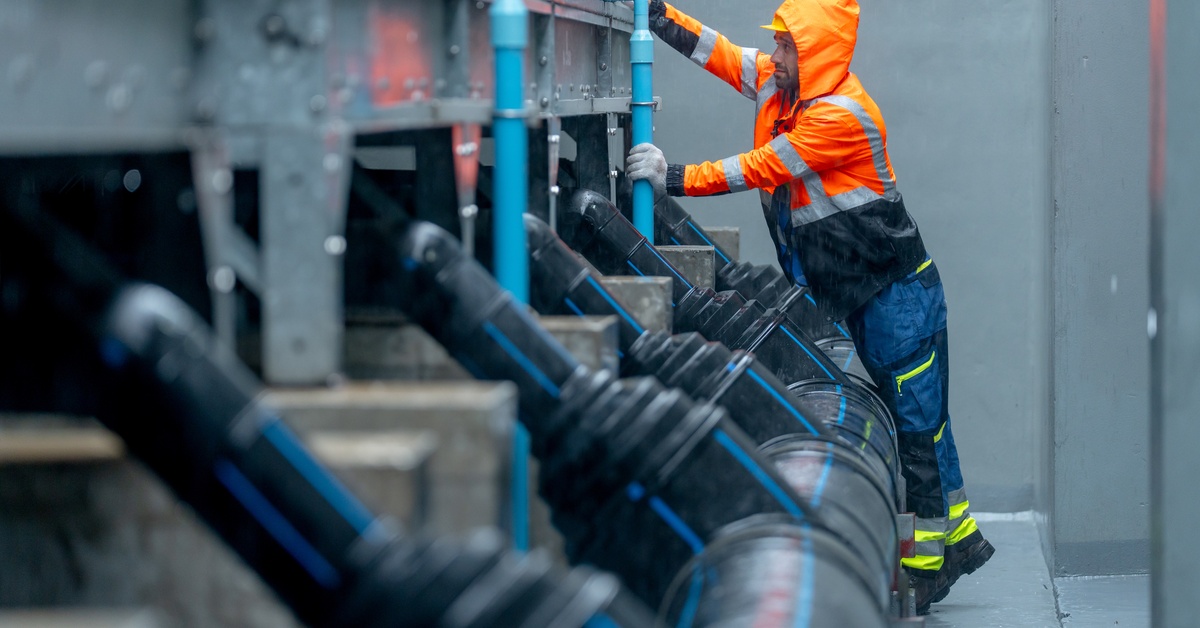 An industrial worker wears flame-resistant rain gear, including a jacket with a hood, while working on pipes outside.