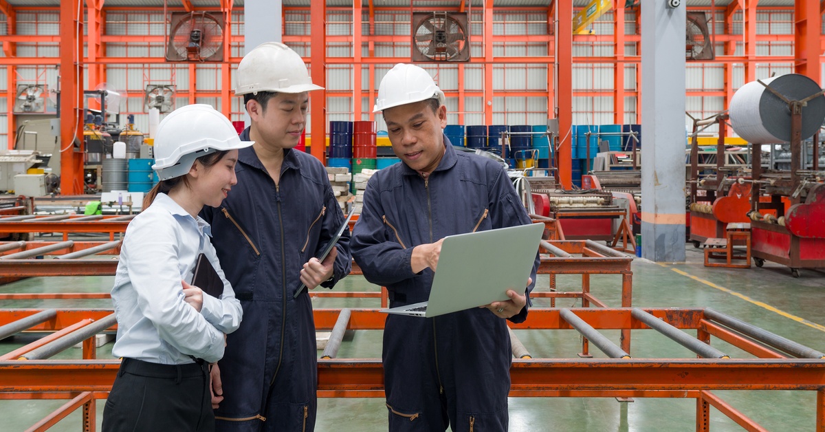 Three industrial workers in white hard hats stand and look at a laptop computer together inside a warehouse environment.