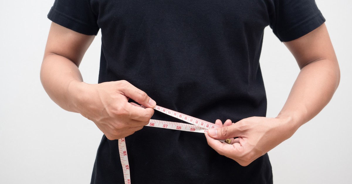 A person in a black T-shirt holds a white tape measure around their waist to take their clothing measurements.