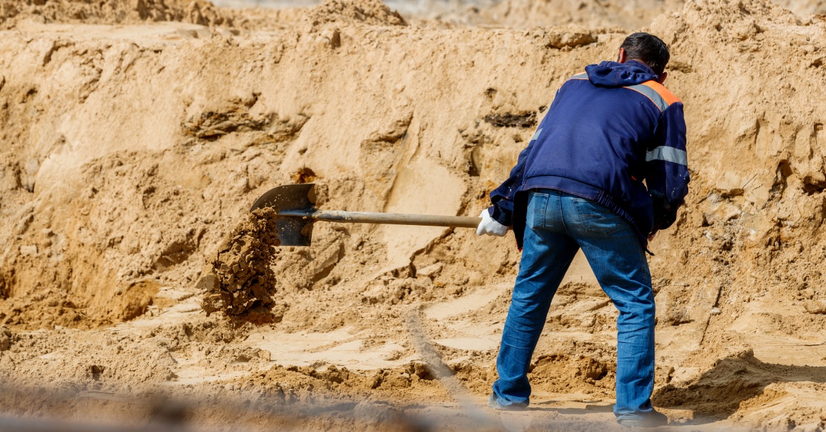 A worker wearing jeans, a blue jacket, and gloves uses a shovel to dig dirt at an outdoor construction site.