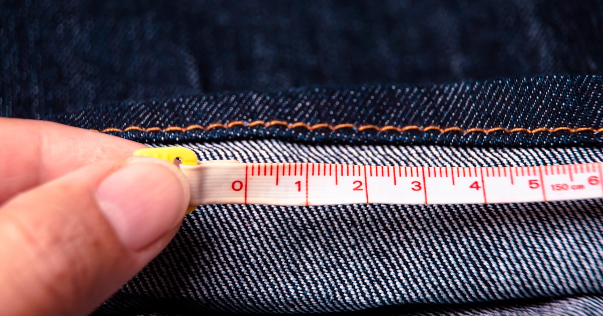 A close-up of a person measuring the bottom hem on a pair of blue jeans with thin white measuring tape.