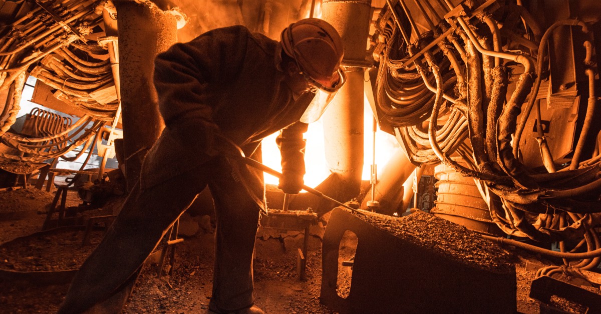An industrial worker in a hard hat stands in front of a fire in a furnace, using a tool to poke at gravel.