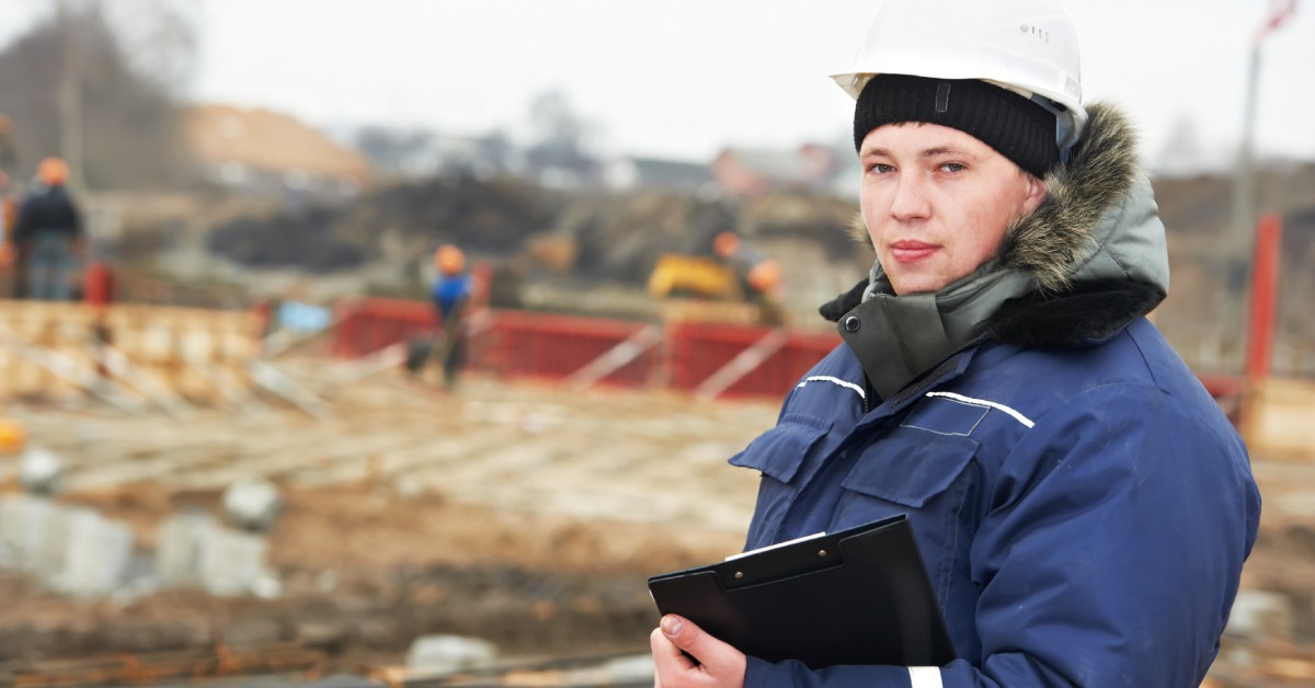 A person in a hooded coat and white hard hat holds a clipboard and stands at an outdoor construction site.