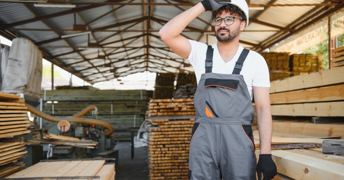 A man wearing overalls, a white hard hat, and black gloves stands surrounded by wooden planks at work.