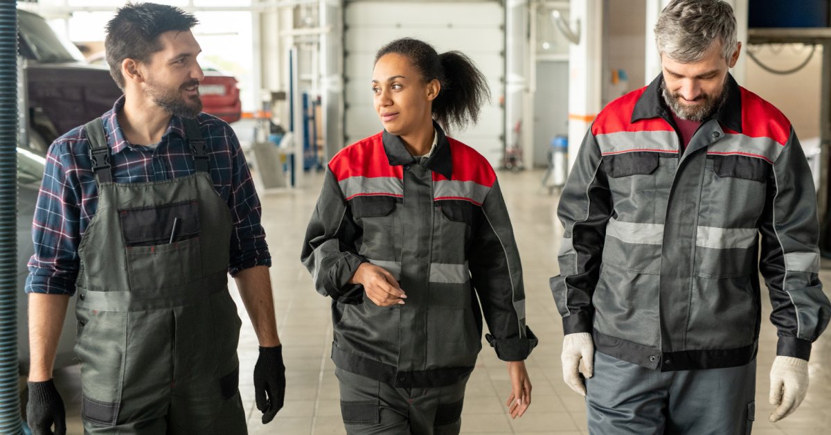 Three workers in industrial work clothes, including one in overalls, walk inside a car garage, looking at each other.